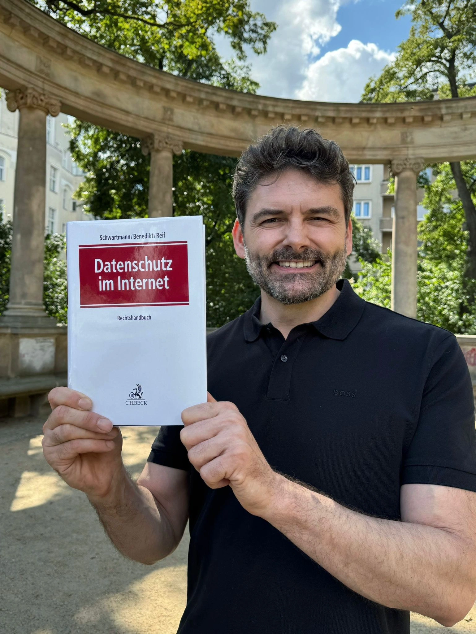 Dr. Thomas Schwenke mit dunklem Poloshirt steht im Freien vor einem klassizistischen Säulenbogen und hält ein Buch hoch auf dessen Deckel oben Schwartmann / Benedikt / Reif steht, darunter in rotem Feld Datenschutz im Internet, darunter Rechts­handbuch und Verlagslogo CH BECK; er lächelt in die Kamera, im Hintergrund Bäume und ein Haus.